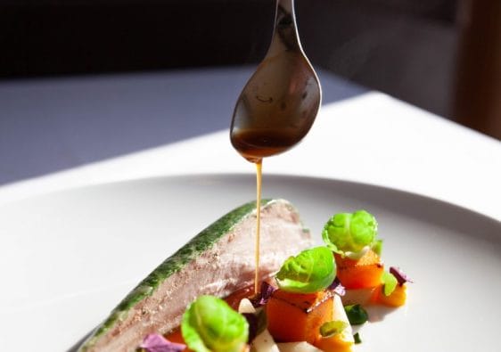 A white plate of Michelin food with a hand dripping sauce over