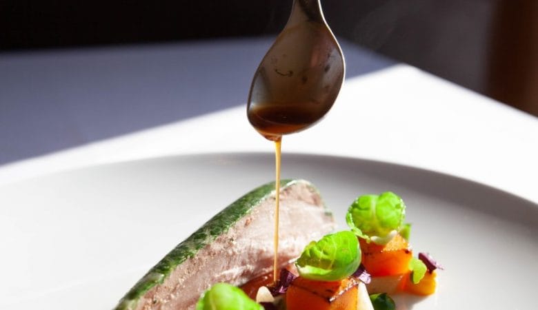 A white plate of Michelin food with a hand dripping sauce over