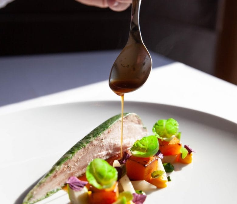 A white plate of Michelin food with a hand dripping sauce over
